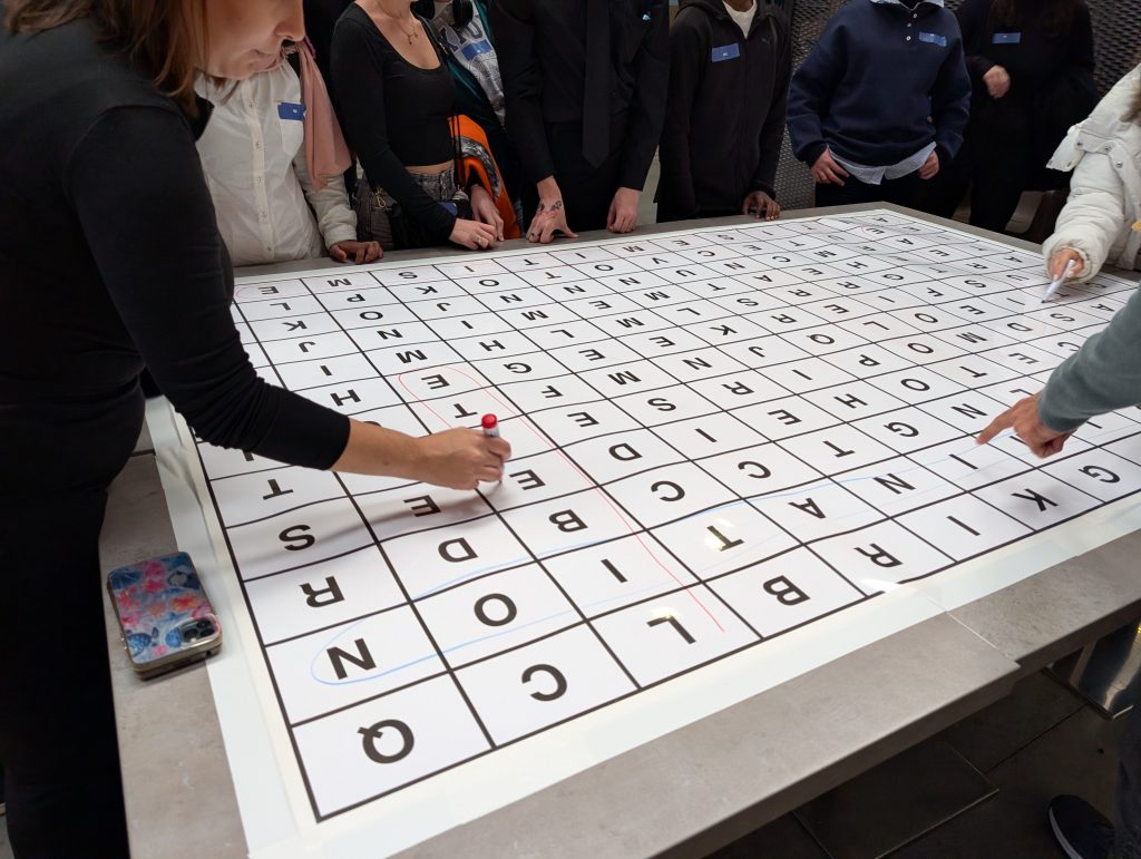 Plusieurs personnes autour d’une grande table avec une grille de lettres, écrivant et pointant des cases au feutre.