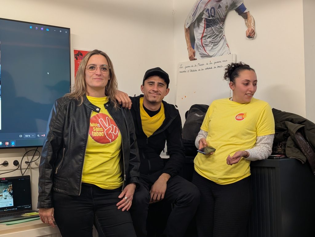 Trois personnes portant des t-shirts jaunes « Mois sans tabac » posent en intérieur, dans une salle équipée d’un écran.
