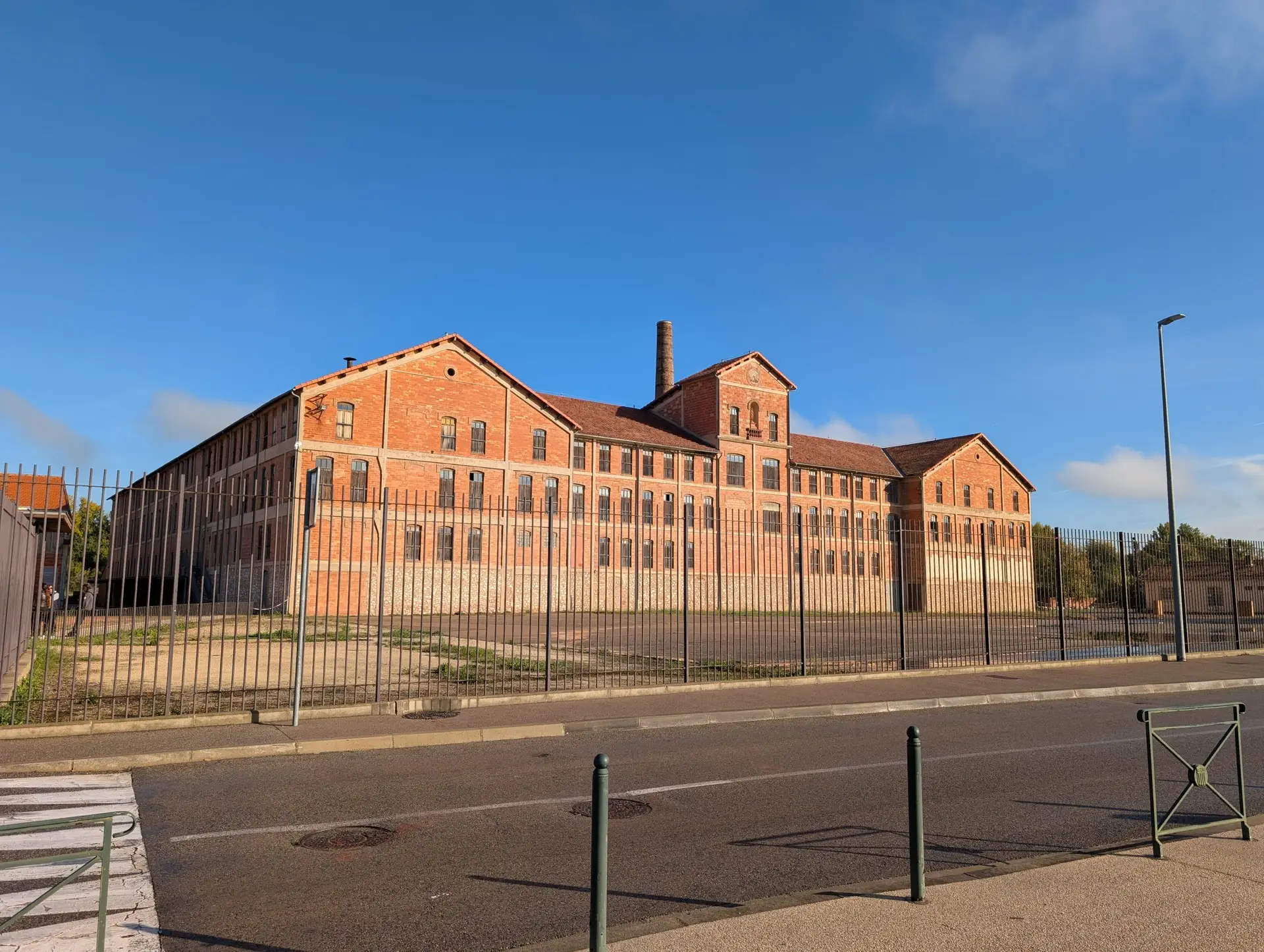Grand bâtiment industriel en briques rouges avec cheminée, derrière une grille, sous un ciel bleu.