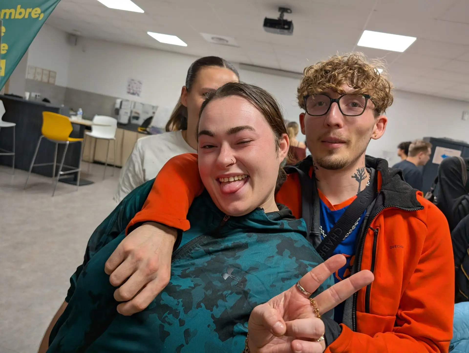 Deux jeunes posent en souriant dans une salle, l’un fait un signe de la paix, avec d’autres personnes en arrière-plan.