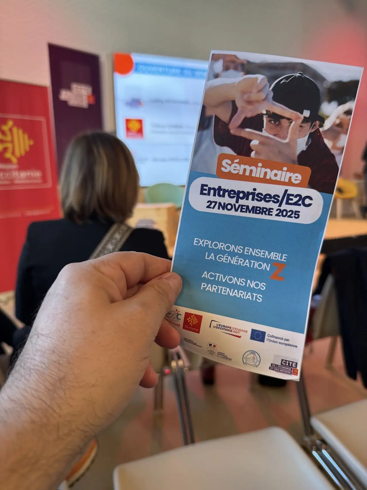 Main tenant un flyer “Séminaire Entreprises/E2C – 27 novembre 2025” dans une salle de conférence, avec écran de présentation en arrière-plan.